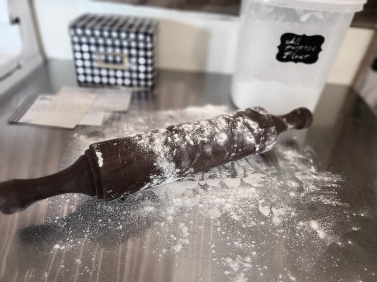 A stainless steel countertop with a rolling pin and flour, set next to a recipe box full of handwritten recipes. This image sets the scene for learning how to cook from scratch.