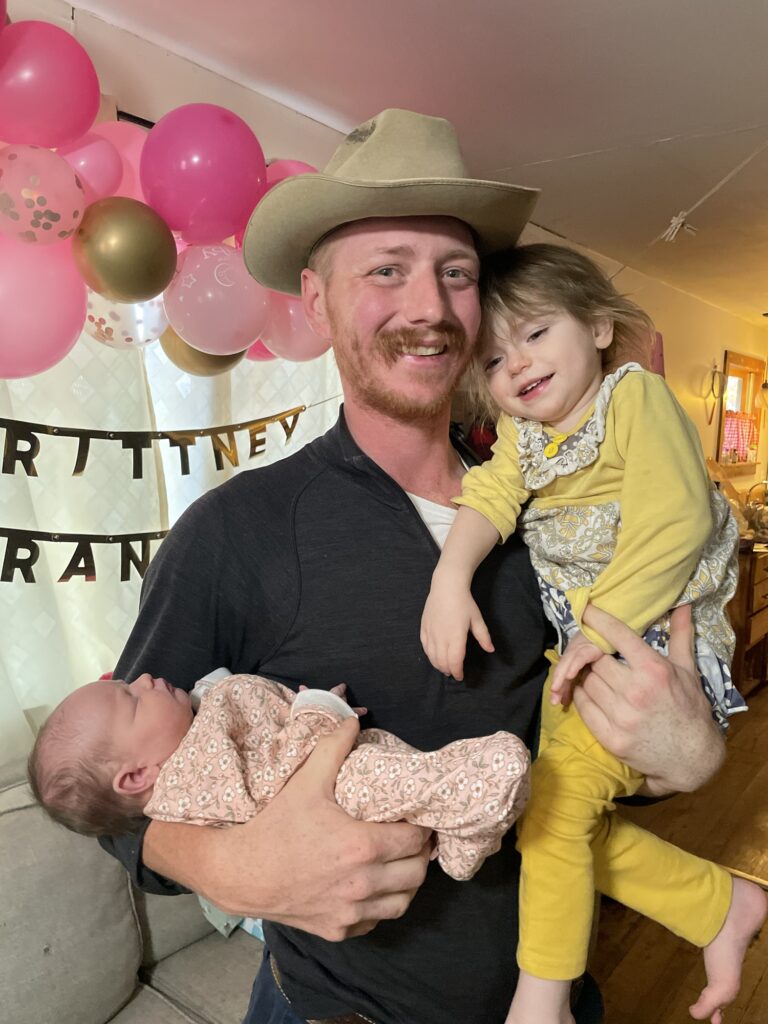 Too Late for the Hospital: My 37-Week Unplanned Home Birth and the Helicopter Ride to the NICU 19 Carson holds his two young daughters, newborn Wrangler and two-year-old Timberlynn, in front of a festive pink and gold balloon and banner display.