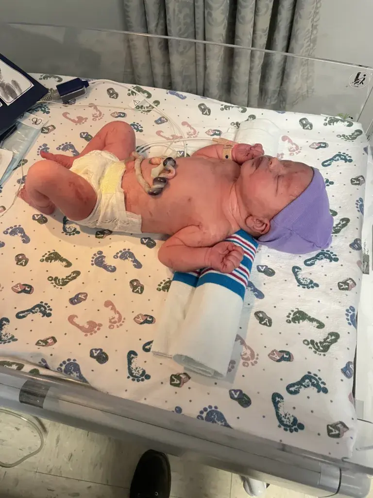 Too Late for the Hospital: My 37-Week Unplanned Home Birth and the Helicopter Ride to the NICU 2 Newborn Wrangler is lying in a warmer with a rolled-up hospital blanket supporting her neck. She still has dried blood visible from her home birth. Her long umbilical cord is coiled on her tummy, and a pulse oximeter is attached to her wrist for monitoring.