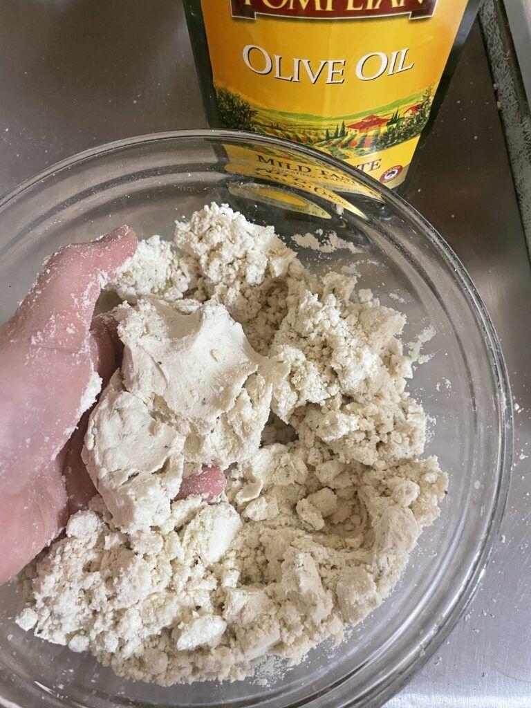 Blue Zone inspired, vegan, garlic rosemary dumpling soup 1 Hand full of dough loosely holding its form over a bowl of dough in the sandy stage forming pea size crumbs.