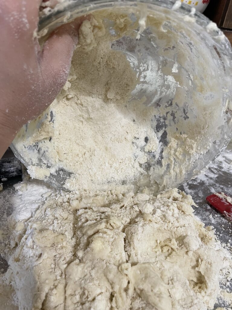 Sourdough biscuit dough is being dumped onto a clean, floured surface with dry bits left over in the bowl to scrape out.