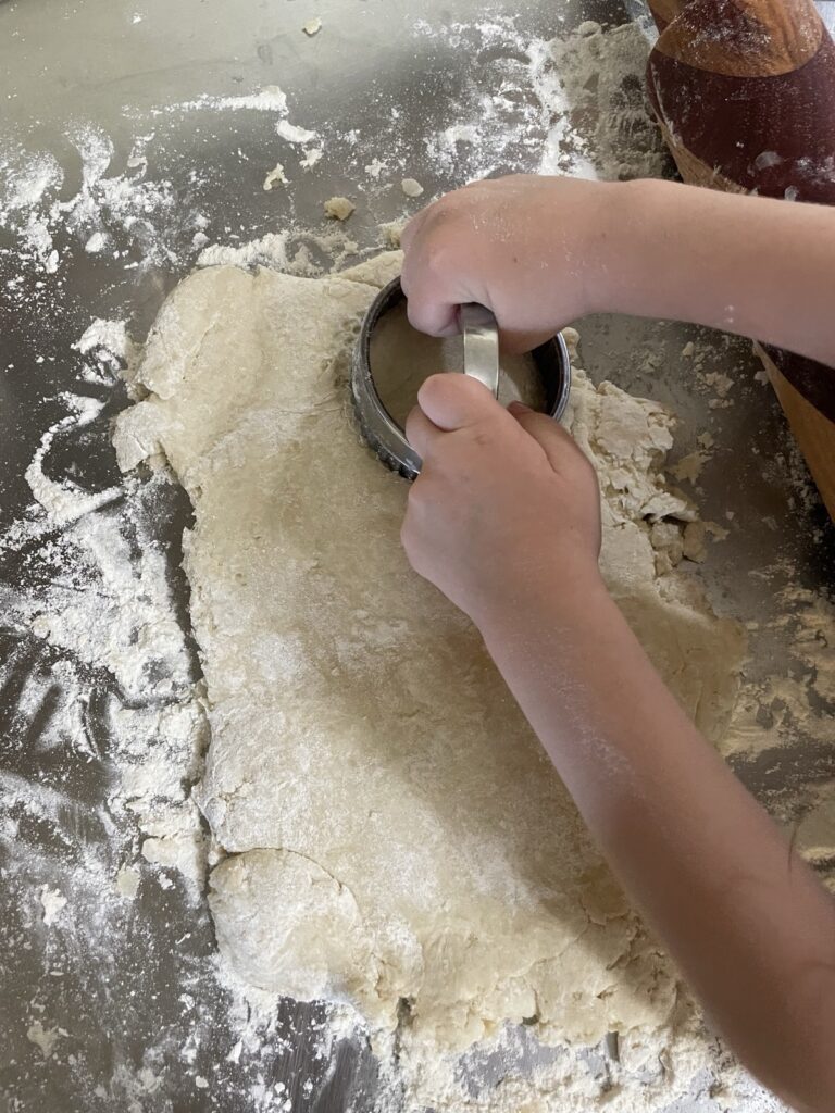 Toddler hands pressing a biscuit cutter straight down into sourdough biscuit dough on a clean, floured workspace.